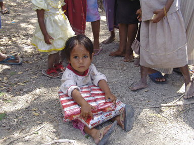 A young child sits on a dirt ground holding a pack of fortified biscuits, surrounded by several other children standing barefoot nearby outdoors.